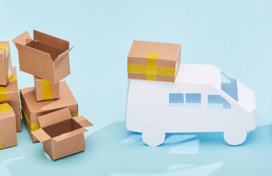 Collection of taped cardboard boxes piled up next to two white toy delivery vans with parcels on their roofs, set against a light blue backdrop.