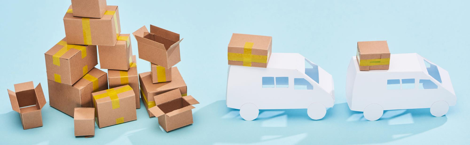Collection of taped cardboard boxes piled up next to two white toy delivery vans with parcels on their roofs, set against a light blue backdrop.