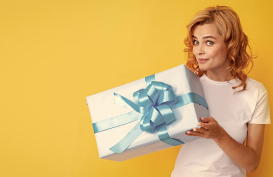 Woman in a white shirt holding a large gift wrapped in light blue ribbon against a yellow background, smiling at the camera.