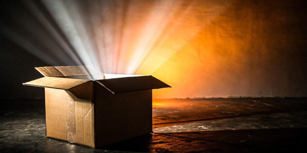 Open cardboard box on a dark floor with a warm orange light spilling out in rays, creating a dramatic glow (informative).