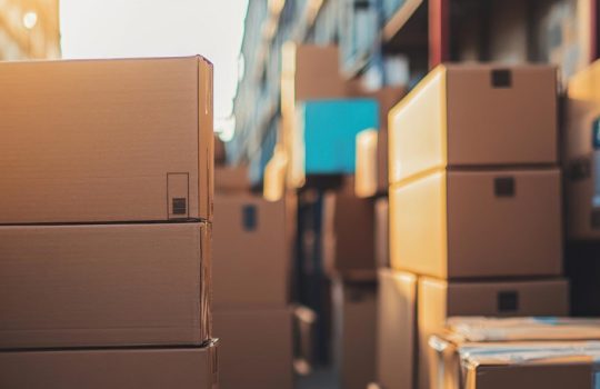 Stack of brown cardboard boxes in a warehouse aisle with warm sunlight on the left side.
